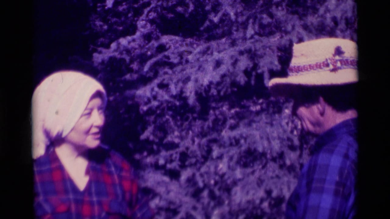 In Agate Springs, Montana, a woman in a white headscarf and red plaid shirt faces a man in a straw hat near evergreen trees in 1975 home-movie footage.