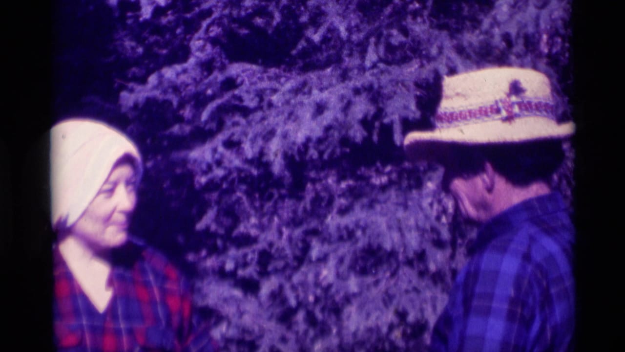 The woman in a white headscarf smiles slightly as she faces the man in a straw hat outdoors in the 1975 Montana home-movie scene.