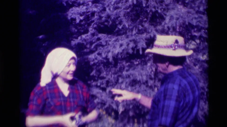 The man in a straw hat gestures toward the woman, who appears to hold a small item, in a candid Agate Springs outdoor exchange from 1975.