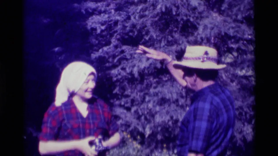 The man raises an arm above shoulder level while facing the woman in a white headscarf, continuing the outdoor neighbor conversation in Montana.