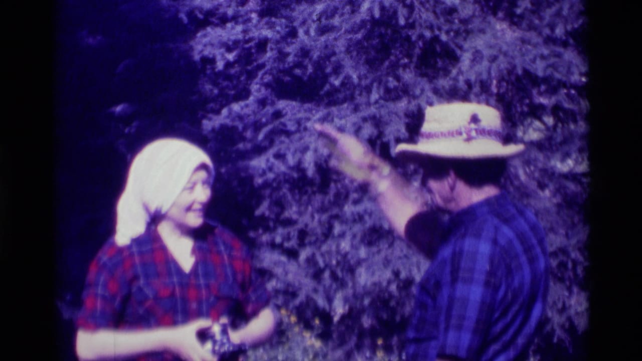 In 1975 Agate Springs home-movie footage, a woman in a white headscarf holds laundry while a man in a straw hat gestures nearby.