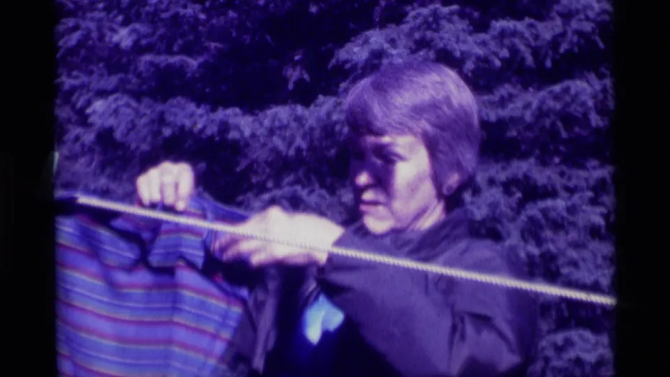 A closer 1975 home-movie frame shows the person fastening a striped wet garment to the backyard clothesline.