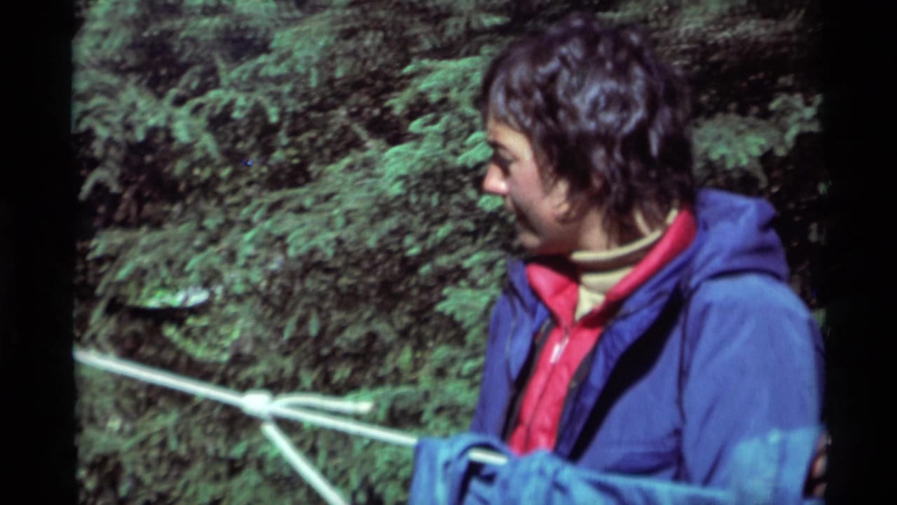 Young man in a blue jacket looks to the side beside white cord and evergreen trees.