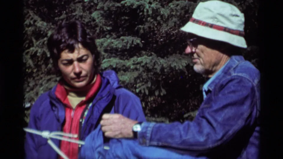 An older man in a light bucket hat and denim jacket joins the younger man as they handle a white rope in the 1975 forest footage.