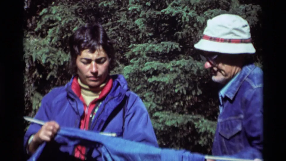 The younger man holds blue fabric or gear while the older man watches beside a white rope in the Agate Springs forest scene.