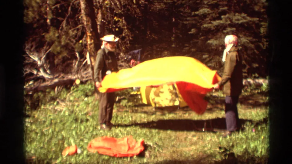 At Agate Springs, Montana in 1975, two adults stand in woods and hold a red and yellow blanket or tarp between them.