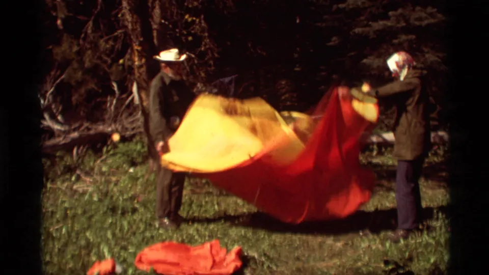 The two adults continue shaking the red and yellow blanket or tarp in a wooded Montana setting on 1975 8mm film.