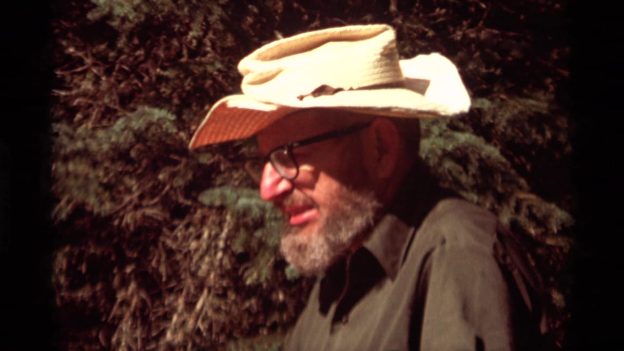 A bearded man in glasses and a light cowboy-style hat sits or crouches near trees in 1975 Montana camping footage.