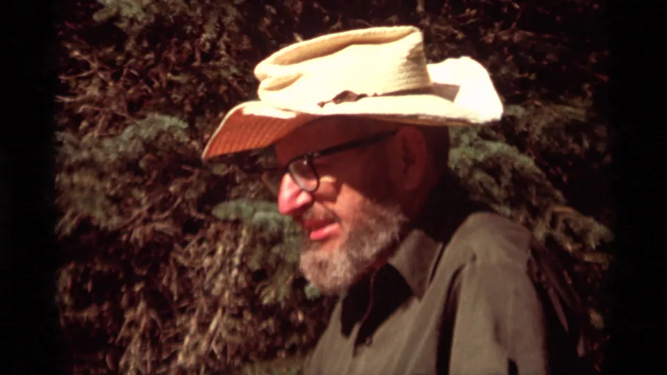 A bearded man in glasses and a light cowboy-style hat sits or crouches near trees in 1975 Montana camping footage.