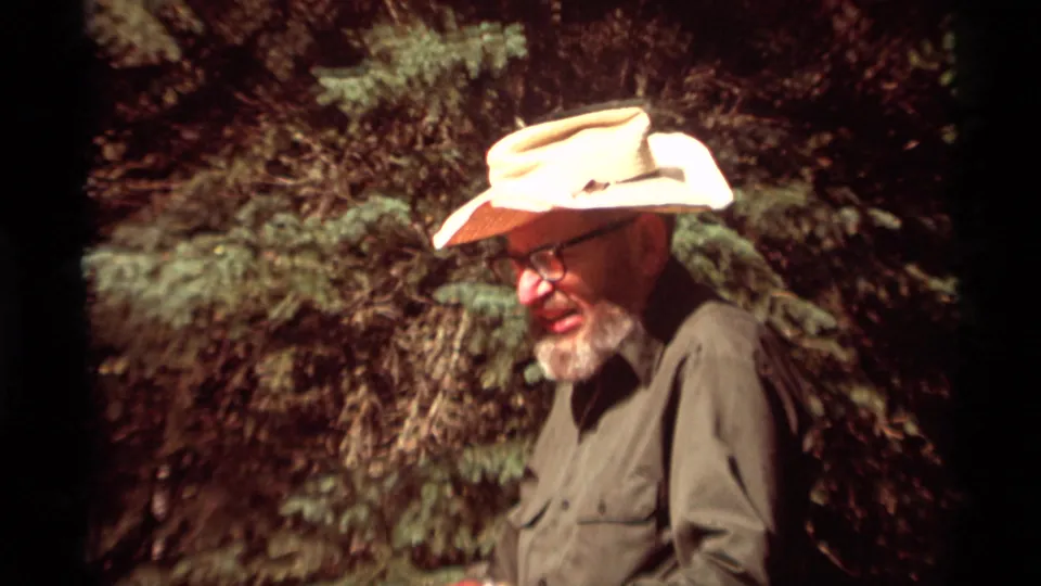 A bearded man wearing glasses and a light cowboy-style hat is framed against evergreens in Agate Springs, Montana, 1975.