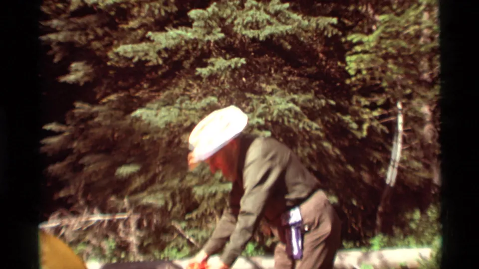 A man in a light hat bends to work with camping gear or tent fabric in a Montana forest clearing in 1975.
