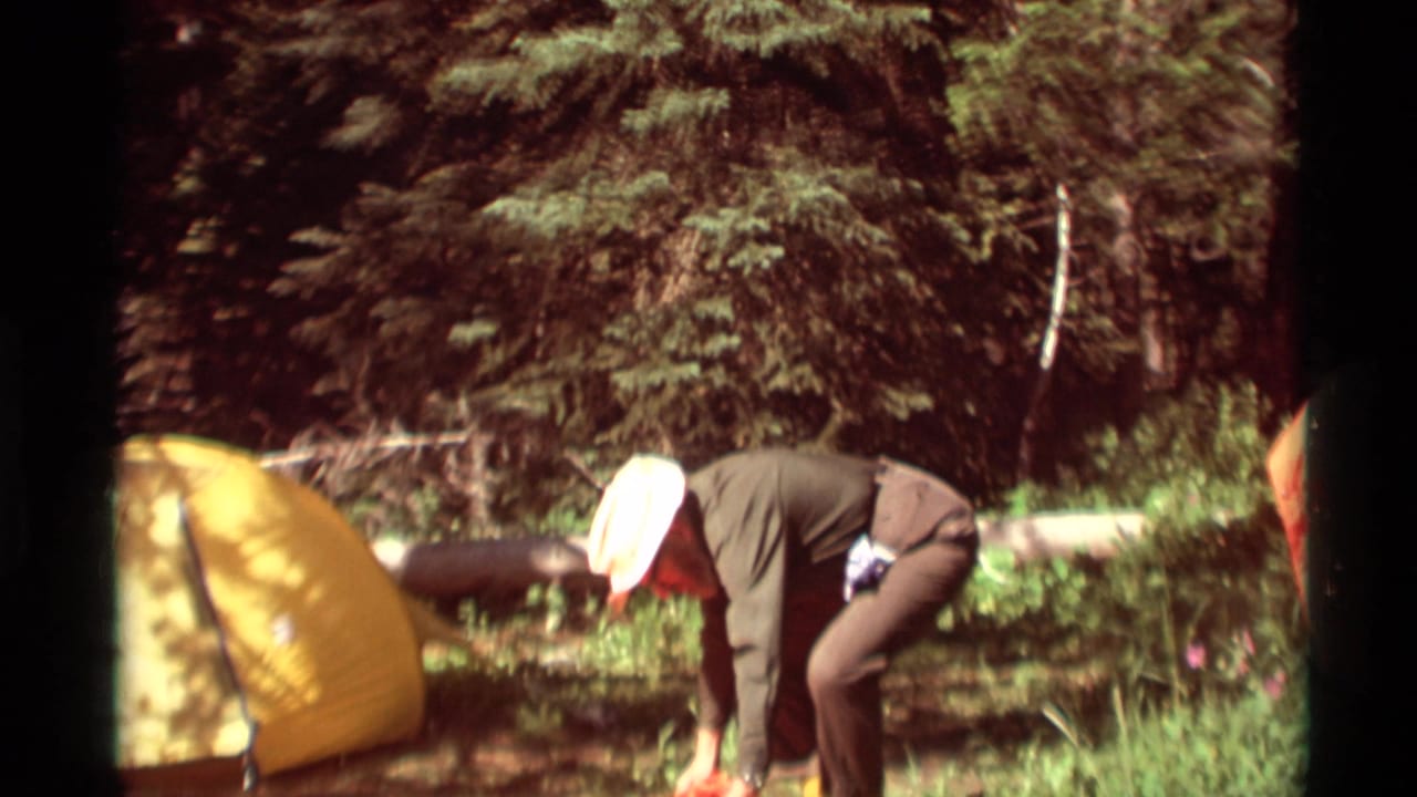 1975 Agate Springs, Montana home-movie frame showing two adults setting up camp near a yellow tent in forest grass.