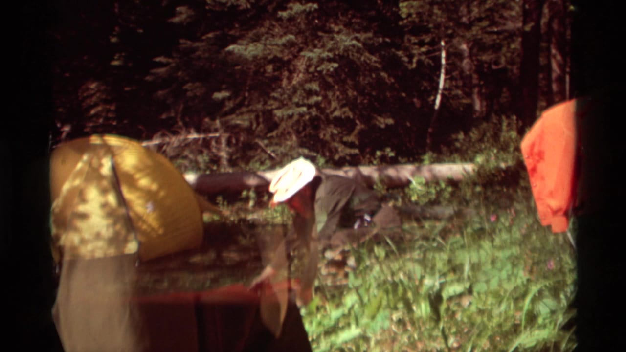 Home-movie frame from the Montana campsite with a yellow tent, one adult bending, and red-orange camping fabric at right.