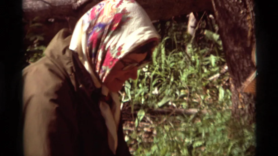 Close home-movie view of a woman in a floral headscarf and glasses helping at the 1975 Montana forest campsite.