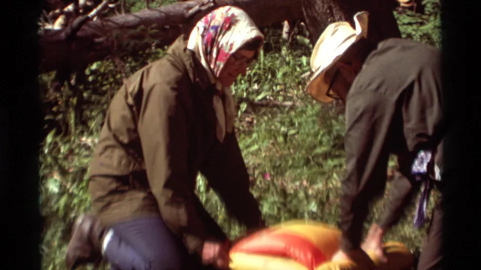 1975 Montana camping frame showing a woman in a headscarf kneeling as another adult helps handle red-orange gear in the forest.