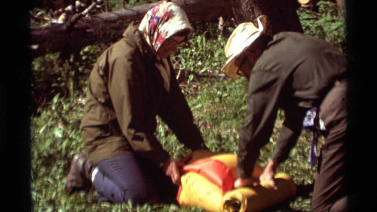Two older adults kneel in a forest while handling bright sleeping bags and camping gear.