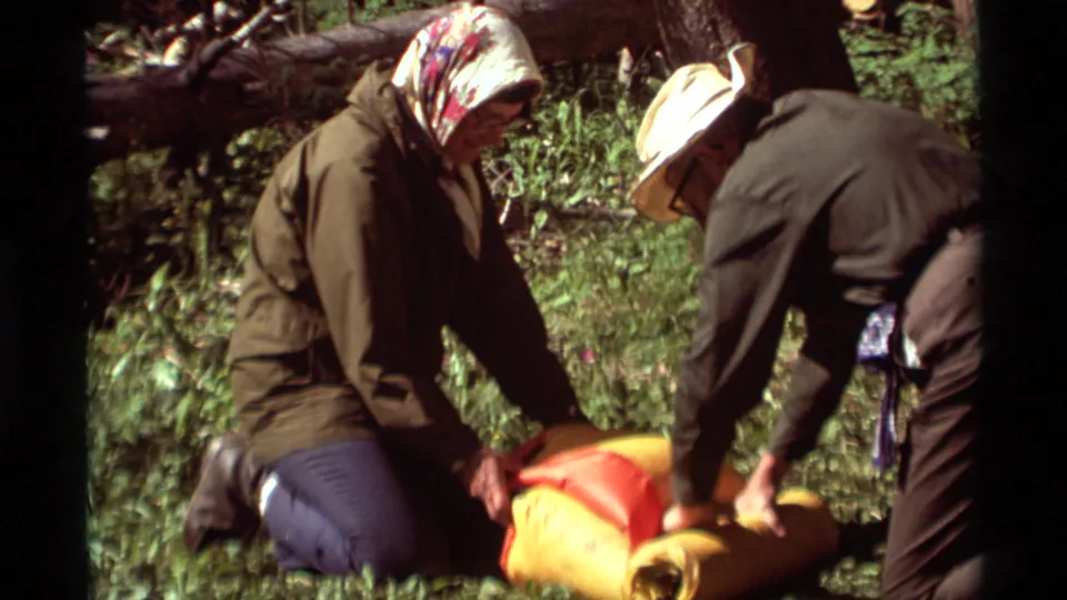 In 1975 Montana home-movie footage, two older adults kneel among trees while arranging orange and yellow sleeping bags for a hike.