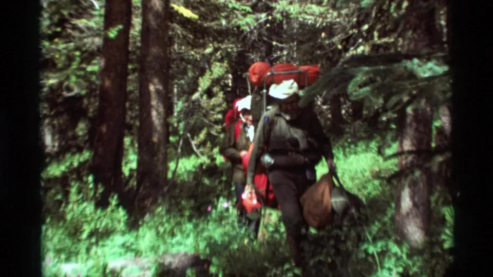 Two older hikers move through dense Montana woods with loaded packs, sleeping bags, and hand-carried gear in 1975 8mm footage.