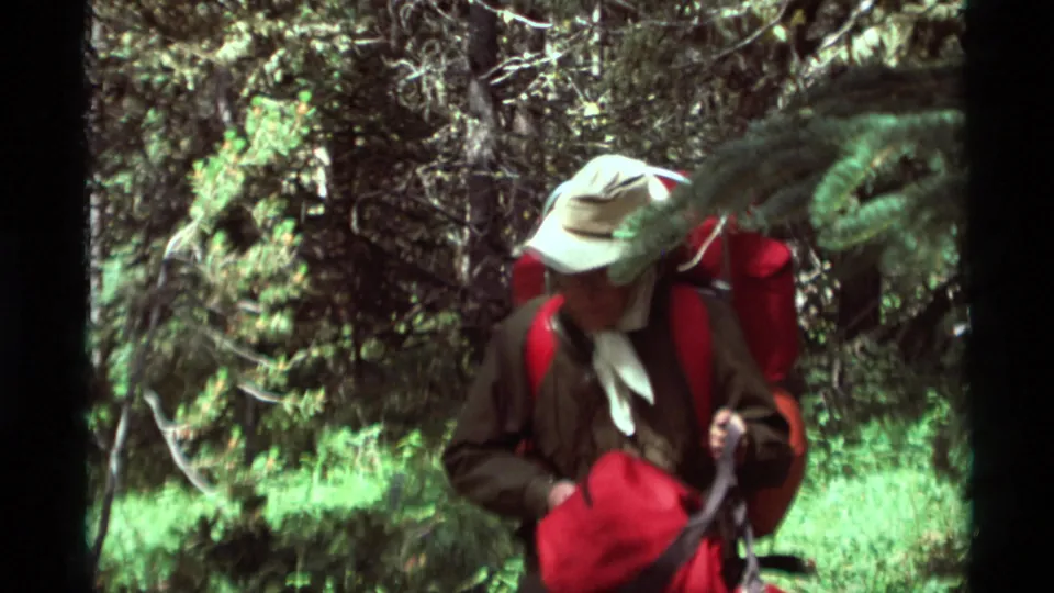Close view of an adult hiker in a light hat and neckerchief carrying red gear through Agate Springs forest footage.