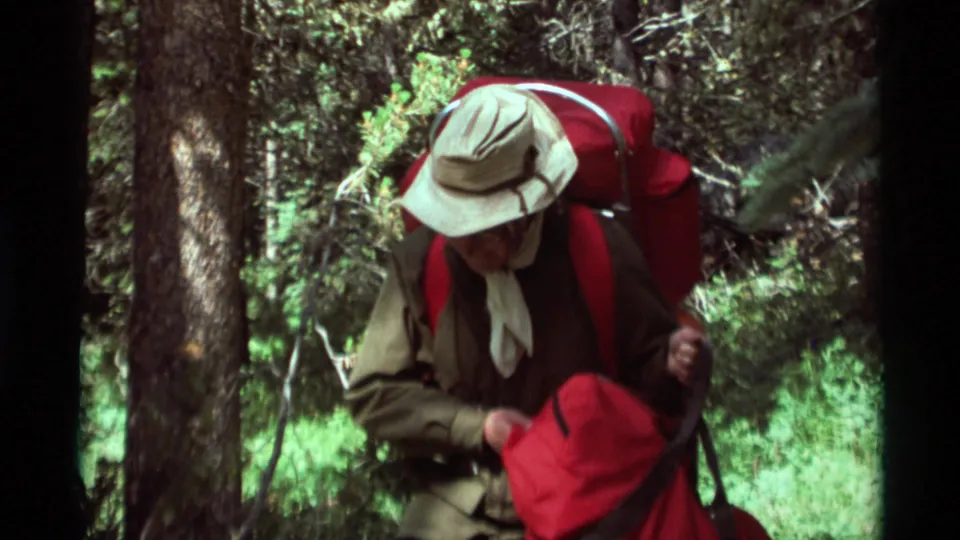 An adult hiker with a light hat, neckerchief, and red backpack moves past trees in 1975 Montana home-movie footage.
