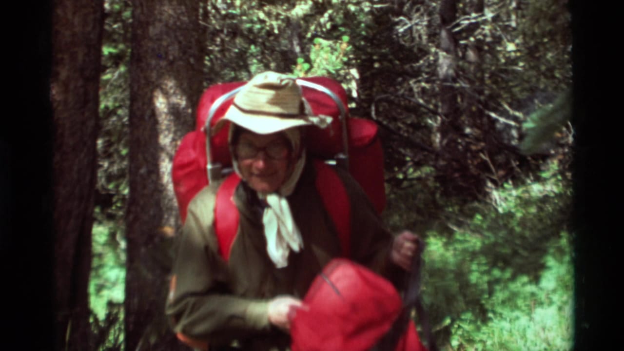 In 1975 Montana home-movie footage, an older woman hikes through dense woods wearing a hat, glasses, and red backpack.