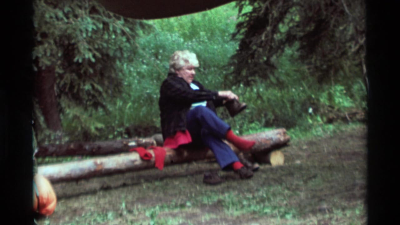 Older woman sitting on cut logs in the woods, lifting one foot near her shoe.