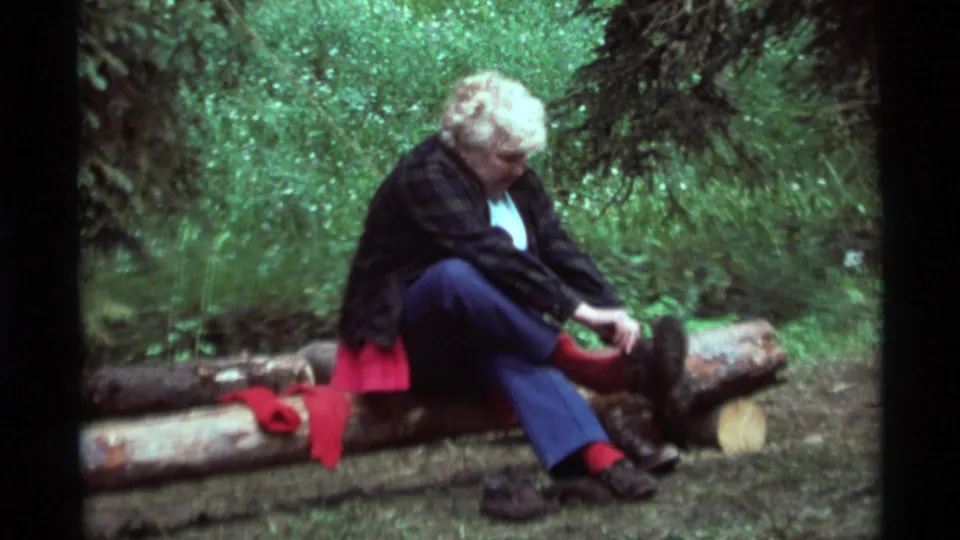 The 1975 home-movie frame shows the woman leaning toward her footwear while seated on cut logs among green trees.