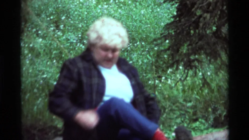 A closer 8mm frame shows the seated woman in the Montana woods, with red socks, dark shoes, and soft green foliage behind her.