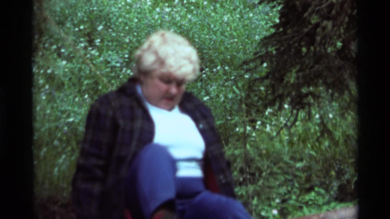 1975 8mm frame from Agate Springs, Montana showing a blonde woman seated near trees and undergrowth.