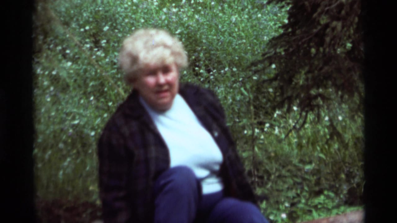 Seated blonde woman looking toward the camera in a wooded green setting.