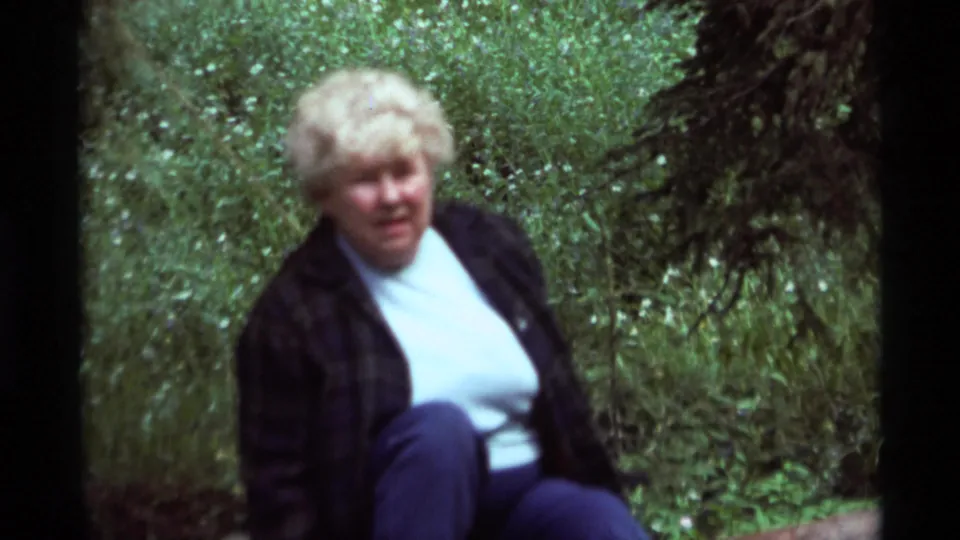 1975 Agate Springs home-movie frame with a seated blonde woman in casual clothing amid foliage.