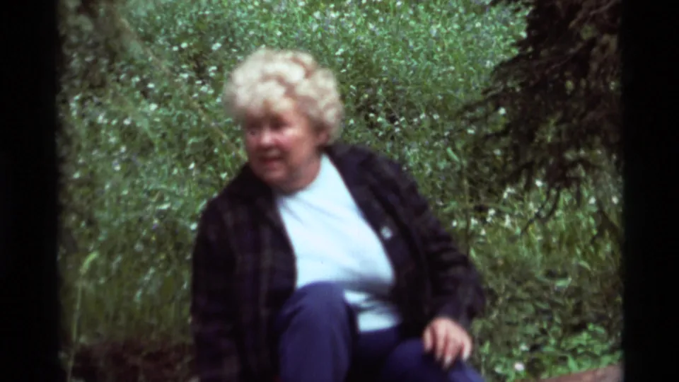1975 8mm outdoor frame showing a seated woman in a plaid jacket and white shirt in Agate Springs, Montana.