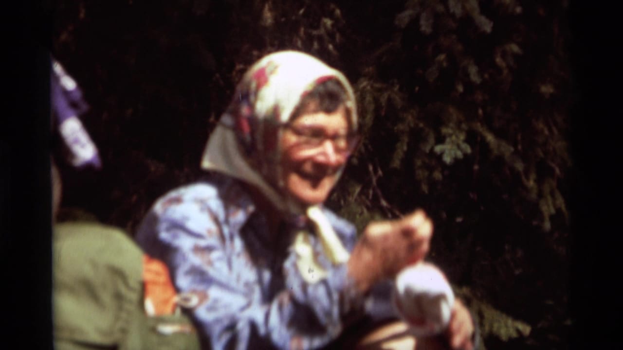 1975 8mm home-movie frame from Agate Springs, Montana, showing a smiling seated woman in a floral headscarf beside outdoor gear.