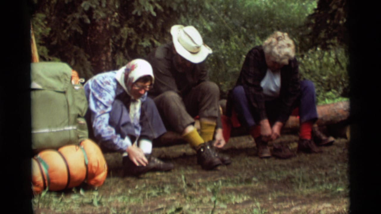 Three adult campers sit on a log beside a green pack and orange sleeping bag.