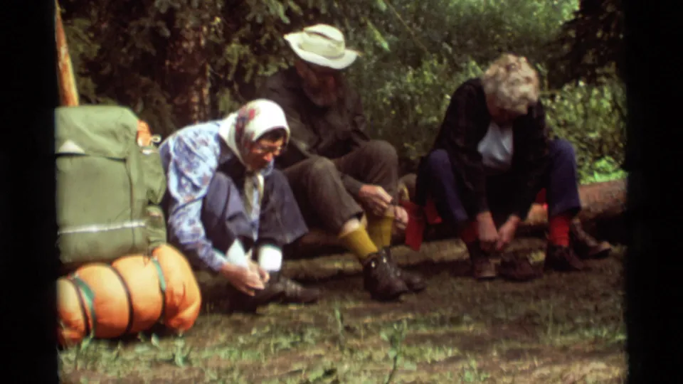 Adult campers in 1970s outdoor clothing sit close together as they adjust or remove footwear during a forest rest stop.