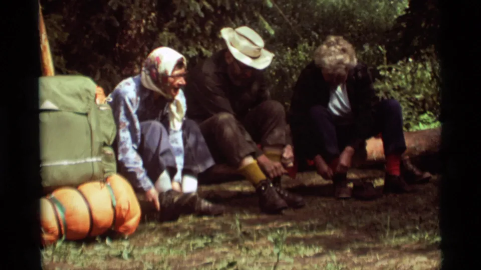 In a wooded Agate Springs rest scene, three adult campers sit on a log while a woman in a headscarf handles her hiking boot.