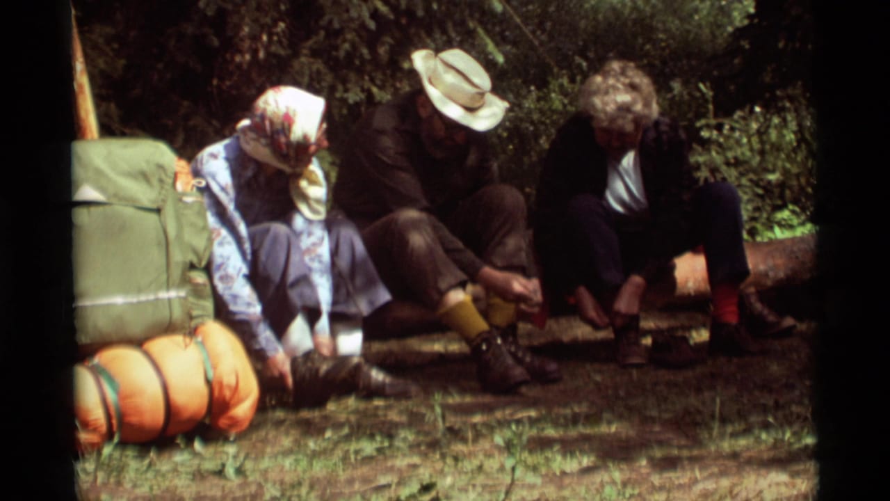 1975 Montana 8mm frame of adults resting beside a green backpack and orange sleeping rolls during a camping stop.