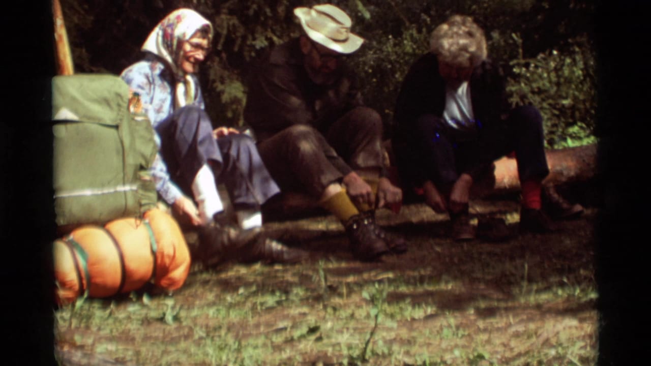 Three adults rest beside a backpack and sleeping rolls while preparing footwear outdoors.