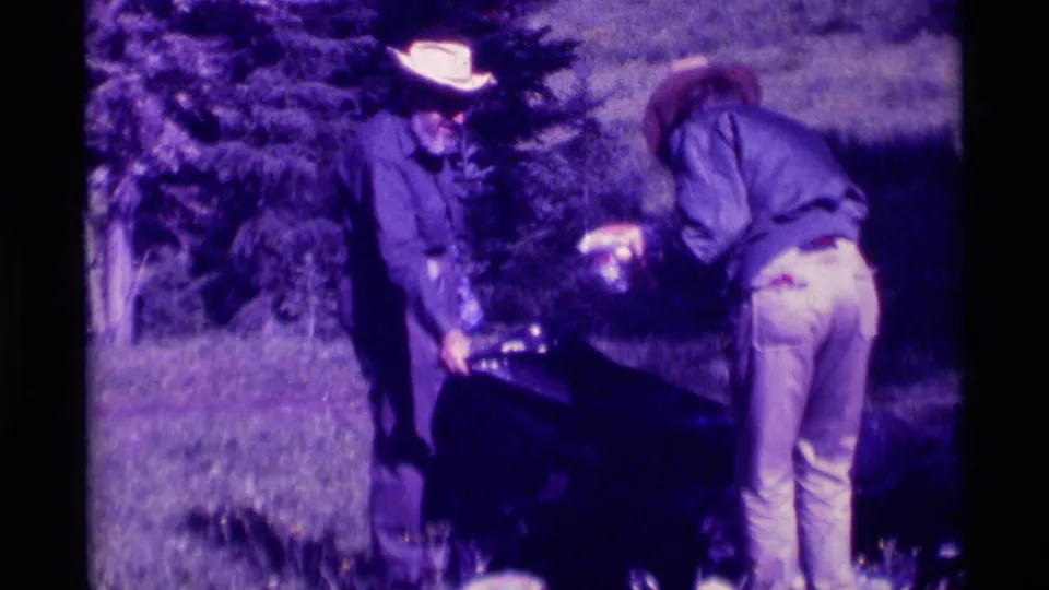 1975 Montana home-movie frame with two adults near a large dark animal or gear shape in an open grassy area.