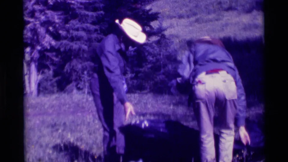 In the 1975 Montana camping footage, two adults bend near a large dark animal or gear shape on grass.