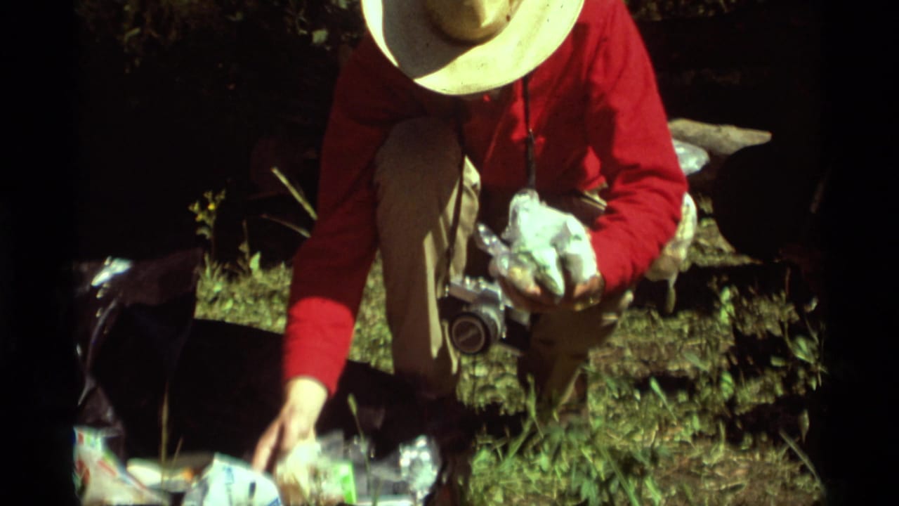 1975 Agate Springs home-movie frame showing a person in a red shirt and straw hat crouched near food and a camera.