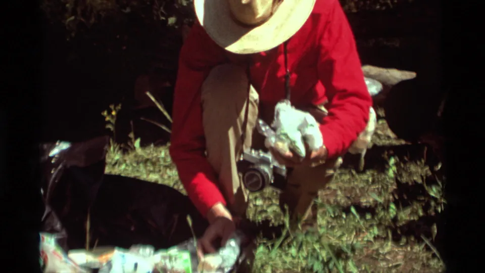 1975 8mm frame from Montana showing a crouching person in a red shirt sorting picnic food on grass near a camera.