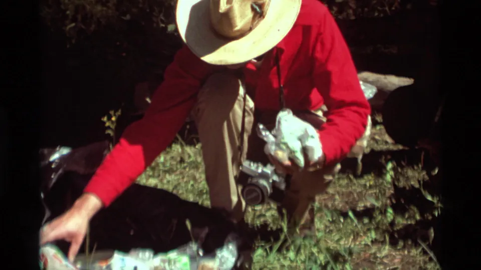 Agate Springs, Montana home-movie frame showing a person in a straw hat reaching over picnic supplies during an outdoor meal setup.