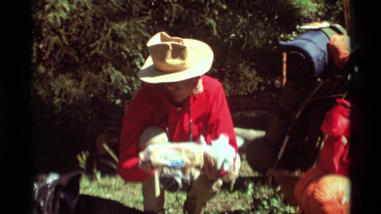 In 1975 Agate Springs, Montana footage, a man in a red shirt and straw hat sits in a wooded area with wrapped bread and gear nearby.