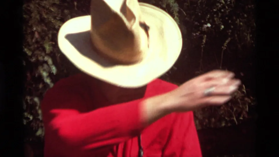 The man in a straw hat and red shirt is framed closely as he raises his hand, with dark trees behind him in the 1975 home movie.