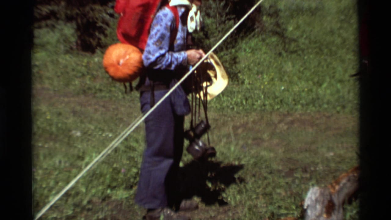 1975 Montana hiking footage shows a standing hiker with a red pack, hat, and dark camera or binocular gear hanging from straps.