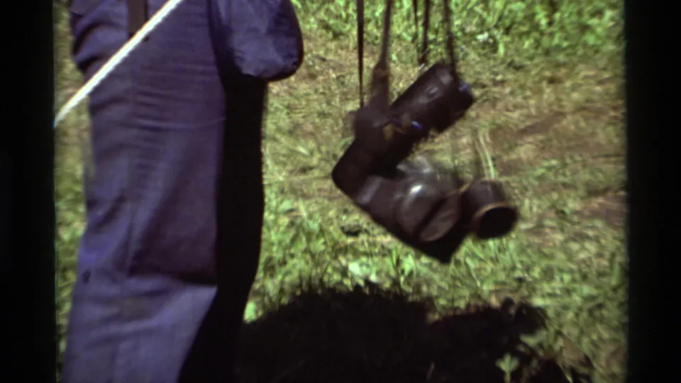 Close 1975 home-movie frame of dark camera or binocular cases swinging from straps beside a hiker in blue trousers.
