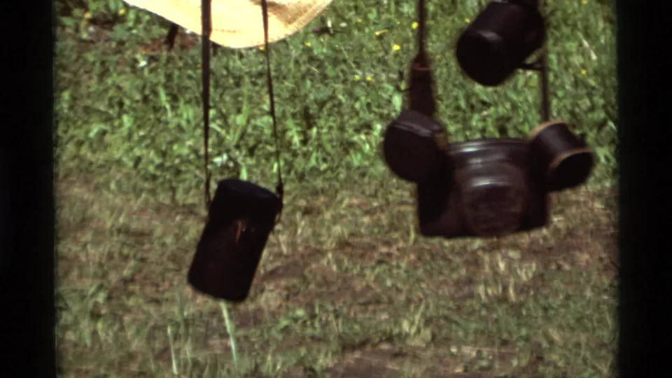 Close 1975 Agate Springs frame showing black camera or binocular cases hanging from straps against a grassy clearing.