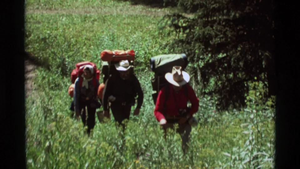 Three travelers with backpacks and hats cross a green forest path in 1975 8mm footage from Agate Springs, Montana.
