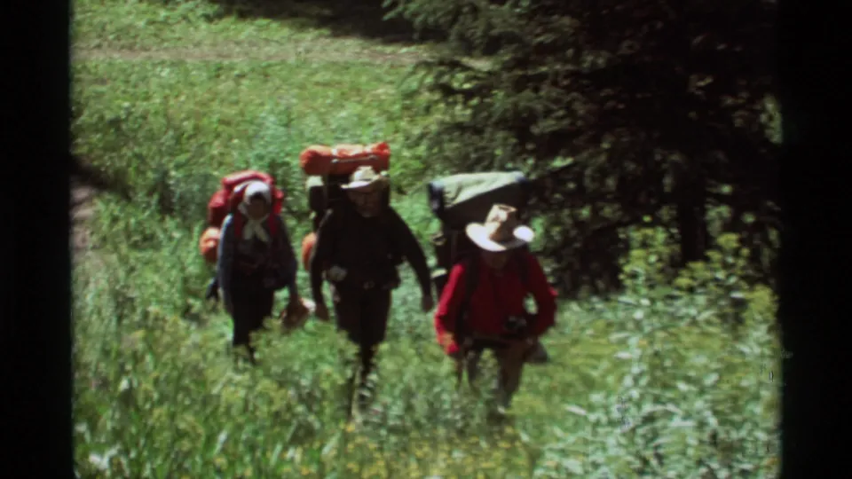 Three backpackers hike through tall green grass in Agate Springs, Montana, in 1975 home-movie footage.